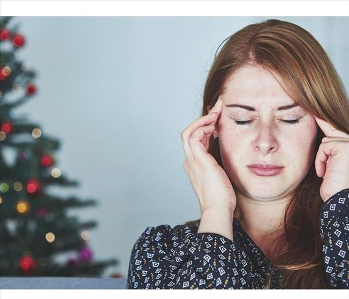 Stressed lady holding hands on her face with eyes closed, wearing sweater with Christmas tree in the background