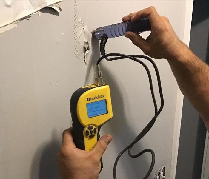Yellow mositure meter reading equipment against a grey wall being used to determine if the sheetrock is wet