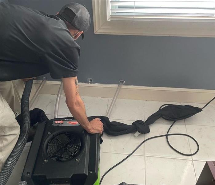 SERVPRO crew member in black shirt and black baseball cap bending down to set air moving equipment after a water damage.